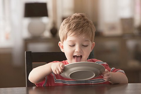 Five-year-old redheaded and adorable boy in a red T-shirt sits at the table and licks a white plate clean with his tongue, which he holds with both hands