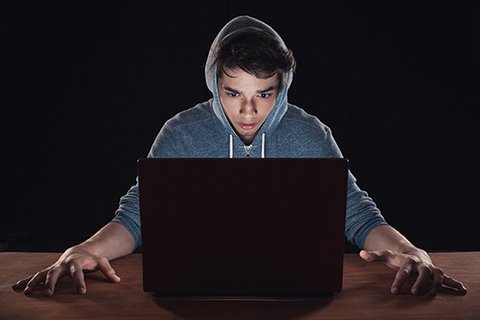 Sechzehnjähriger Jugendlicher mit Kapuzenpulli sitzt erschrocken und gefesselt in ein Laptop starrend am Tisch