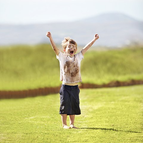 Five-year-old adorable rascal barefoot on a freshly mown meadow cheers with stretched arms and clenched fists, with completely dirty face, dirty T-shirt and low-hanging short