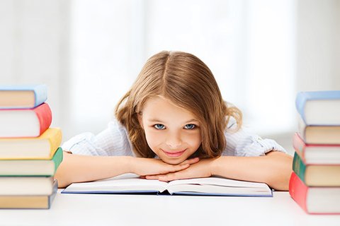 Achtjähriges Mädchen mit hellbraunen langen Haaren liegt entspannt lächelnd auf aufgeklapptem Schulbuch, auf ordentlichem, strukturierten Schreibtisch