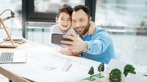 Vater im Homeoffice mit seinem Sohn macht ein Selfie am Schreibtisch