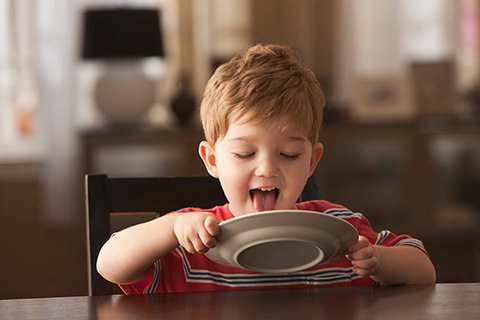 Fünfjähriger rothaariger und liebenswerter Junge im roten T-Shirt sitzt am Tisch und schleckt mit der Zunge einen weißen Teller sauber, den er mit beiden Händen hält