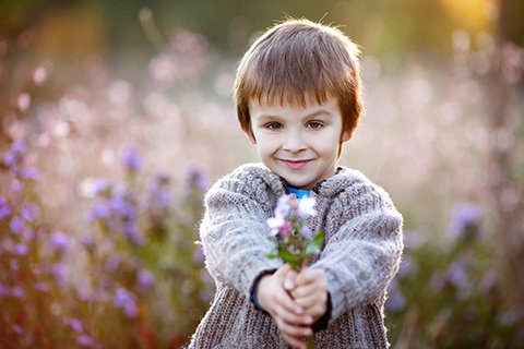 Sechsjähriger blonder Junge mit grauer Strickweste stehend vor Blumenwiese, streckt einen selbstgepflückten Blumenstrauß entgegen