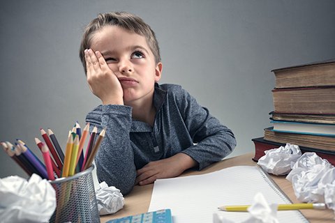 Desperate seven-year-old boy with his head resting, looking to the ceiling, at a chaotic desk between balls of paper, books and pens.